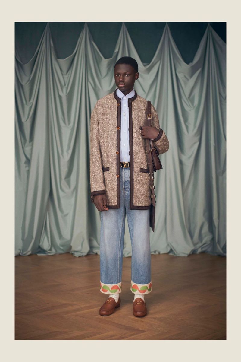 An African American man poses in a tweed jacket, light-wash jeans, and loafers, with a shoulder bag, against a draped backdrop.
