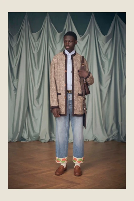 An African American man poses in a tweed jacket, light-wash jeans, and loafers, with a shoulder bag, against a draped backdrop.