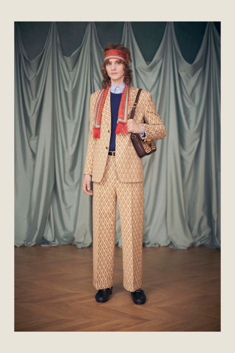 Full shot of a male model wearing a patterned tan suit, scarf, and headband, posing in a studio with a grey curtain backdrop.