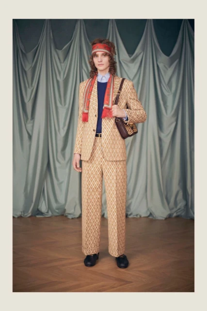 Full shot of a male model wearing a patterned tan suit, scarf, and headband, posing in a studio with a grey curtain backdrop.