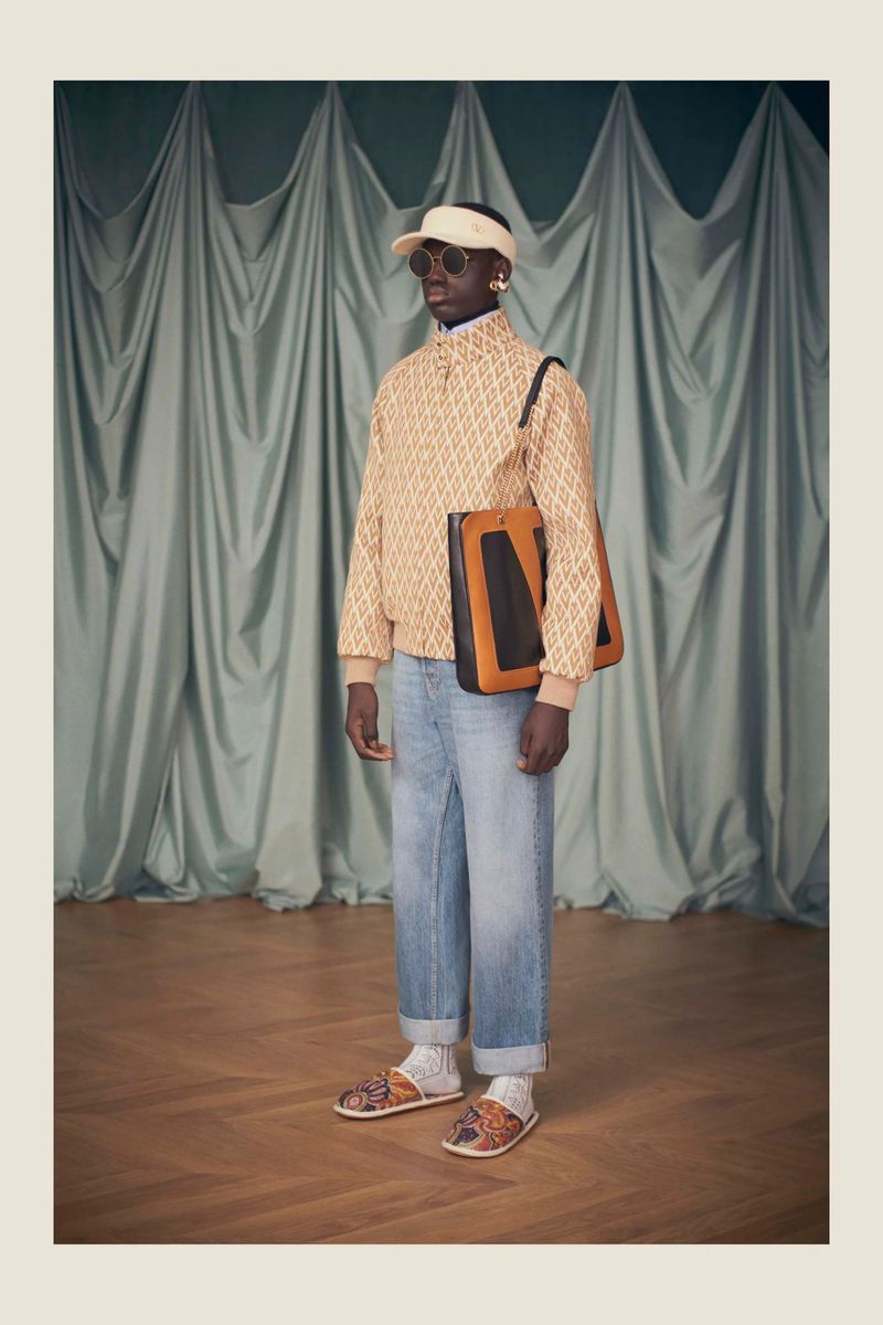 A dark-skinned male model poses in a light patterned jacket, blue jeans, visor, and sandals, carrying a bag against a draped curtain.