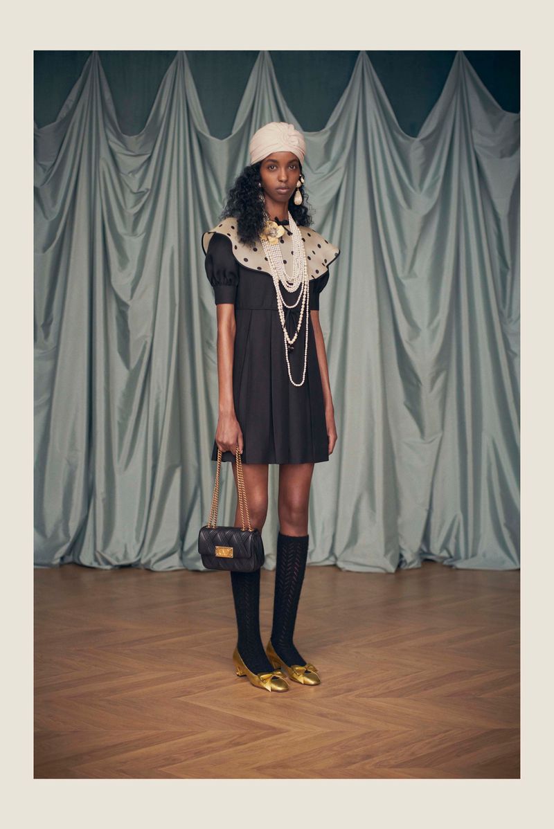 A stylish woman in a black dress, accessorized with pearls, a black purse, gold shoes, and a cream-colored turban, poses elegantly.