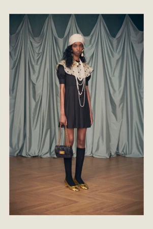 A stylish woman in a black dress, accessorized with pearls, a black purse, gold shoes, and a cream-colored turban, poses elegantly.