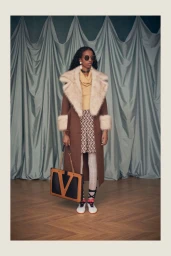 Model standing in a brown fur-trimmed coat holding a large Valentino logo tote bag in front of a draped curtain backdrop.