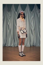 Model in a cream checked mini dress, round sunglasses, and headscarf wearing a crossbody bag in front of draped curtains.