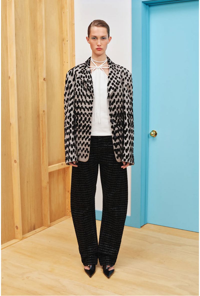 A woman in a speckled blazer and wide-legged black pants stands in front of a blue door and wooden paneling in a studio setting.