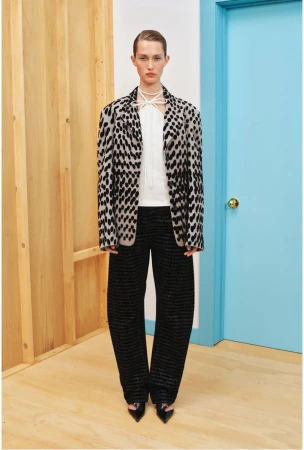 A woman in a speckled blazer and wide-legged black pants stands in front of a blue door and wooden paneling in a studio setting.
