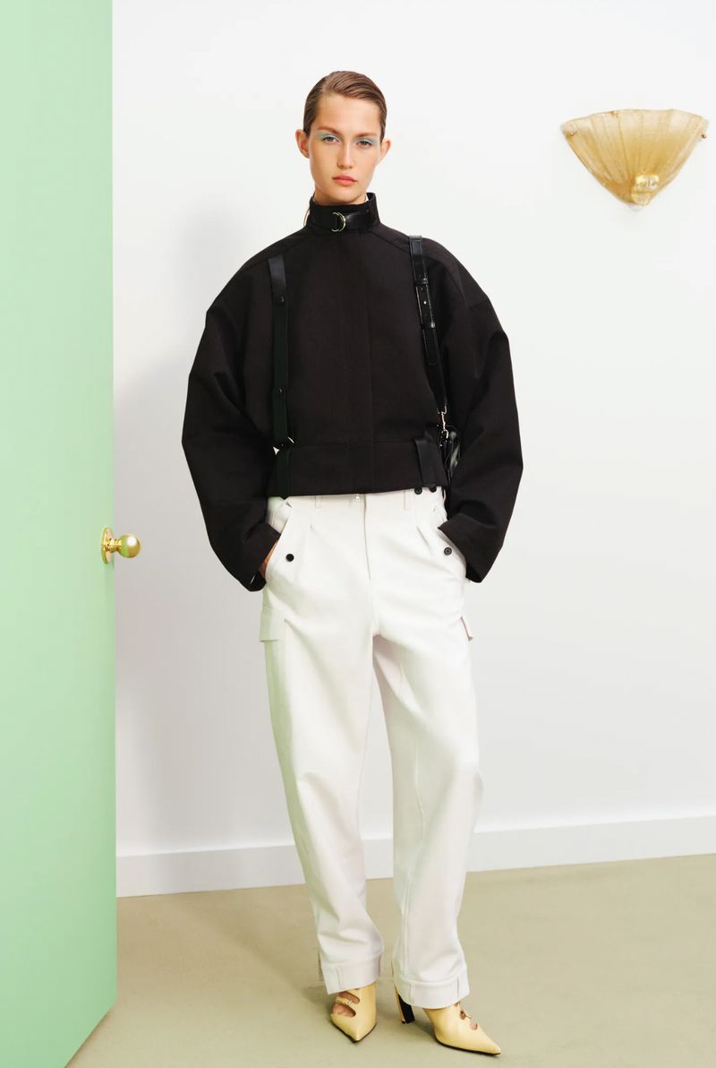Fashion model poses indoors wearing a black high-necked sweatshirt, white trousers, and yellow heels. Bright lighting and simple backdrop.