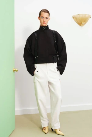 Fashion model poses indoors wearing a black high-necked sweatshirt, white trousers, and yellow heels. Bright lighting and simple backdrop.