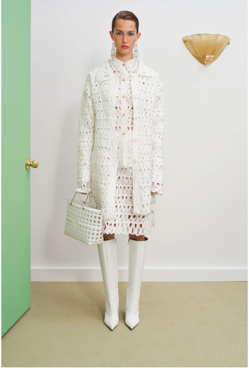 A woman models a white eyelet coat, dress, knee-high boots, and purse in front of a door and light fixture in a studio.