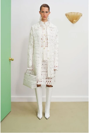 A woman models a white eyelet coat, dress, knee-high boots, and purse in front of a door and light fixture in a studio.