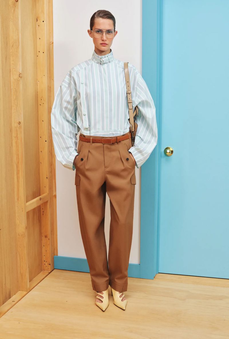 Woman in retro-inspired fashion wearing a striped shirt, brown trousers, and neutral heels against a split-color wall indoors.