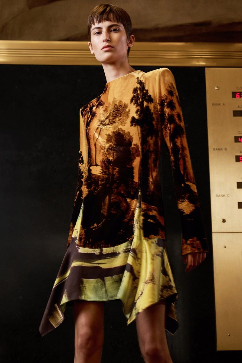 A young woman in a long-sleeved, patterned minidress stands indoors against a dark background with gold trim in a fashion shot.