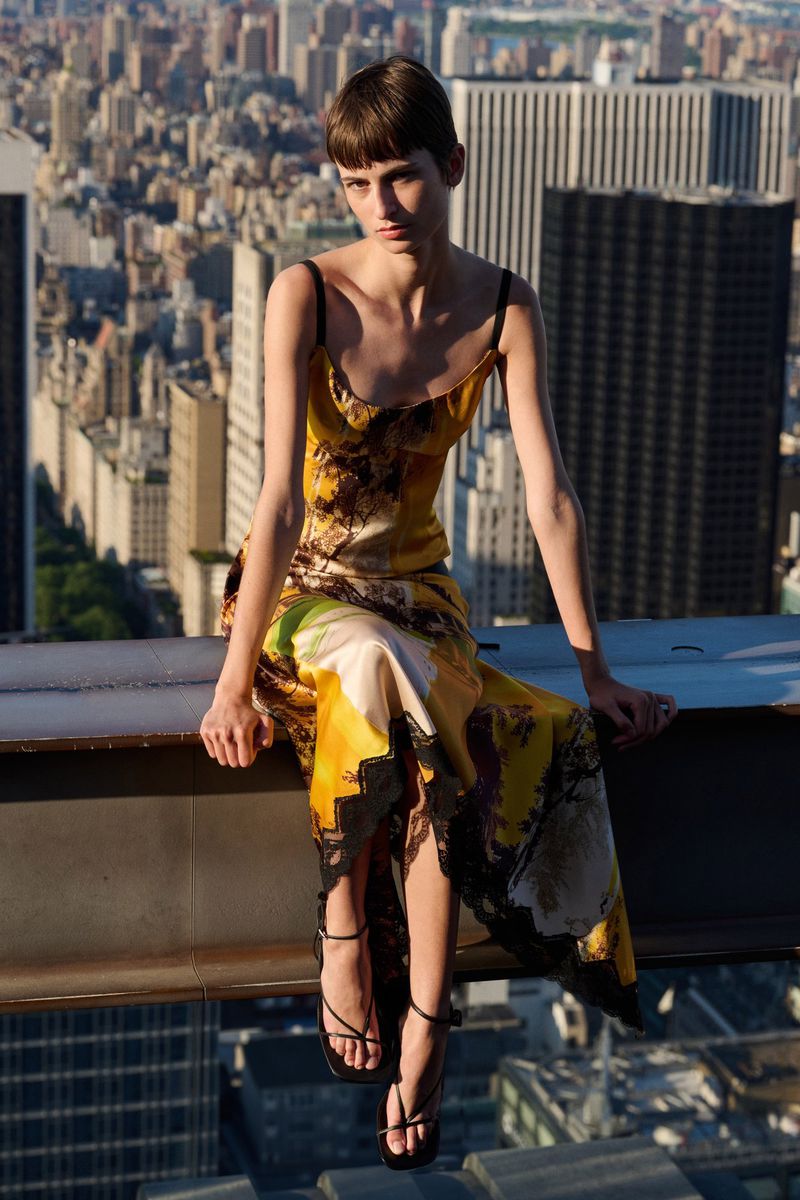 A woman in a patterned dress sits on a metal beam against a city skyline backdrop, showcasing fashion in an urban environment.