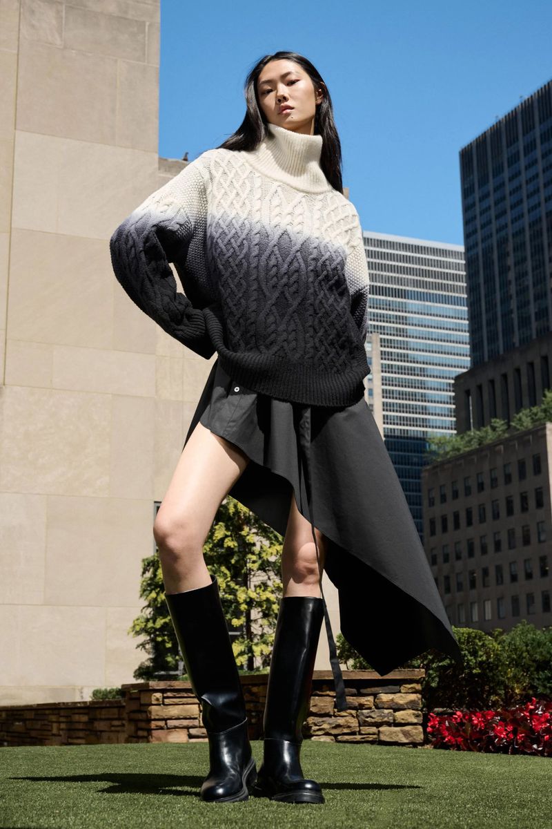 Fashionable young woman in stylish sweater, black skirt, and boots poses against urban backdrop of modern city buildings and blue sky.