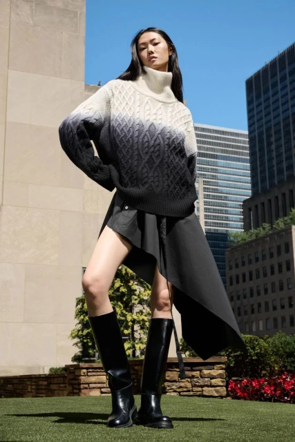 Fashionable young woman in stylish sweater, black skirt, and boots poses against urban backdrop of modern city buildings and blue sky.