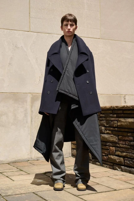 An adult fashion model poses in a full-length shot, wearing a stylish navy blue coat, grey shirt, and pants against a neutral urban backdrop.