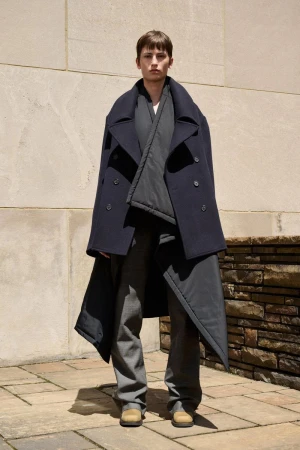 An adult fashion model poses in a full-length shot, wearing a stylish navy blue coat, grey shirt, and pants against a neutral urban backdrop.