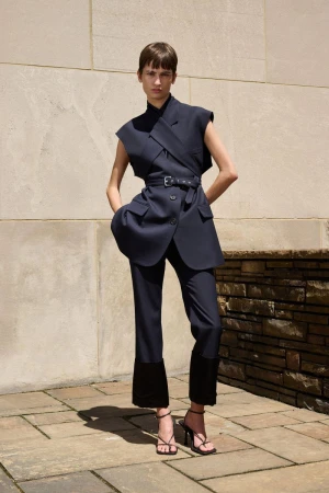 Model posing outdoors in a navy sleeveless belted tailored suit and strappy heels against a stone wall for MONSE Resort 2025.