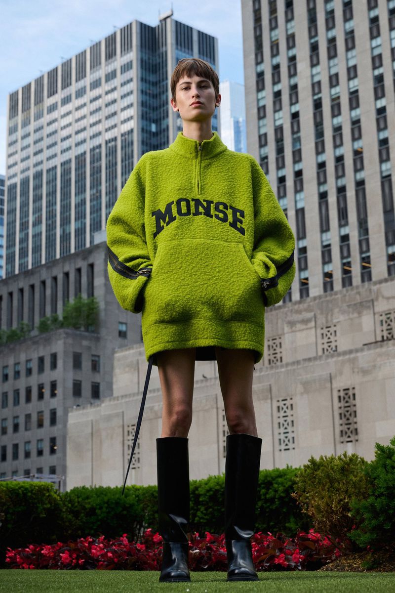 Fashion model in oversized green fleece sweatshirt with MONSE logo and knee-high boots stands against a backdrop of city buildings.