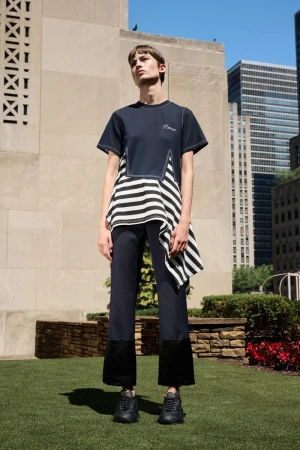 Model wearing an asymmetrical striped navy top and cropped trousers standing on a lawn against city buildings.