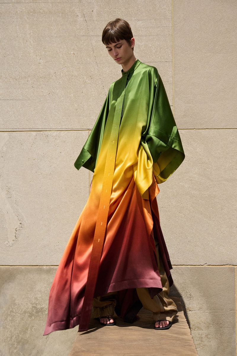 Fashion shot of a person wearing a long, flowing, ombre-colored robe in shades of green, yellow, orange, and pink on a wooden platform.