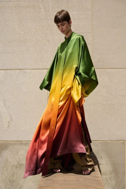 Fashion shot of a person wearing a long, flowing, ombre-colored robe in shades of green, yellow, orange, and pink on a wooden platform.
