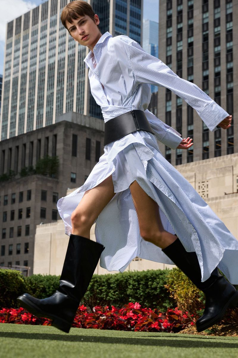 Model Maud van der Meet strides in a blue striped shirt dress, black corset belt, and tall black boots against city skyscrapers.