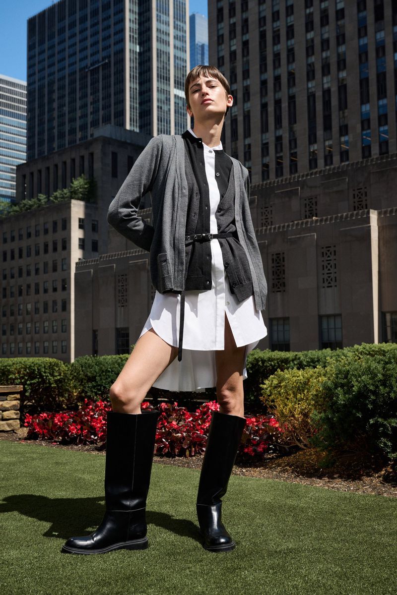 Fashionable woman in black boots, wearing a cardigan and shirtdress, poses outdoors in front of modern architecture and greenery.
