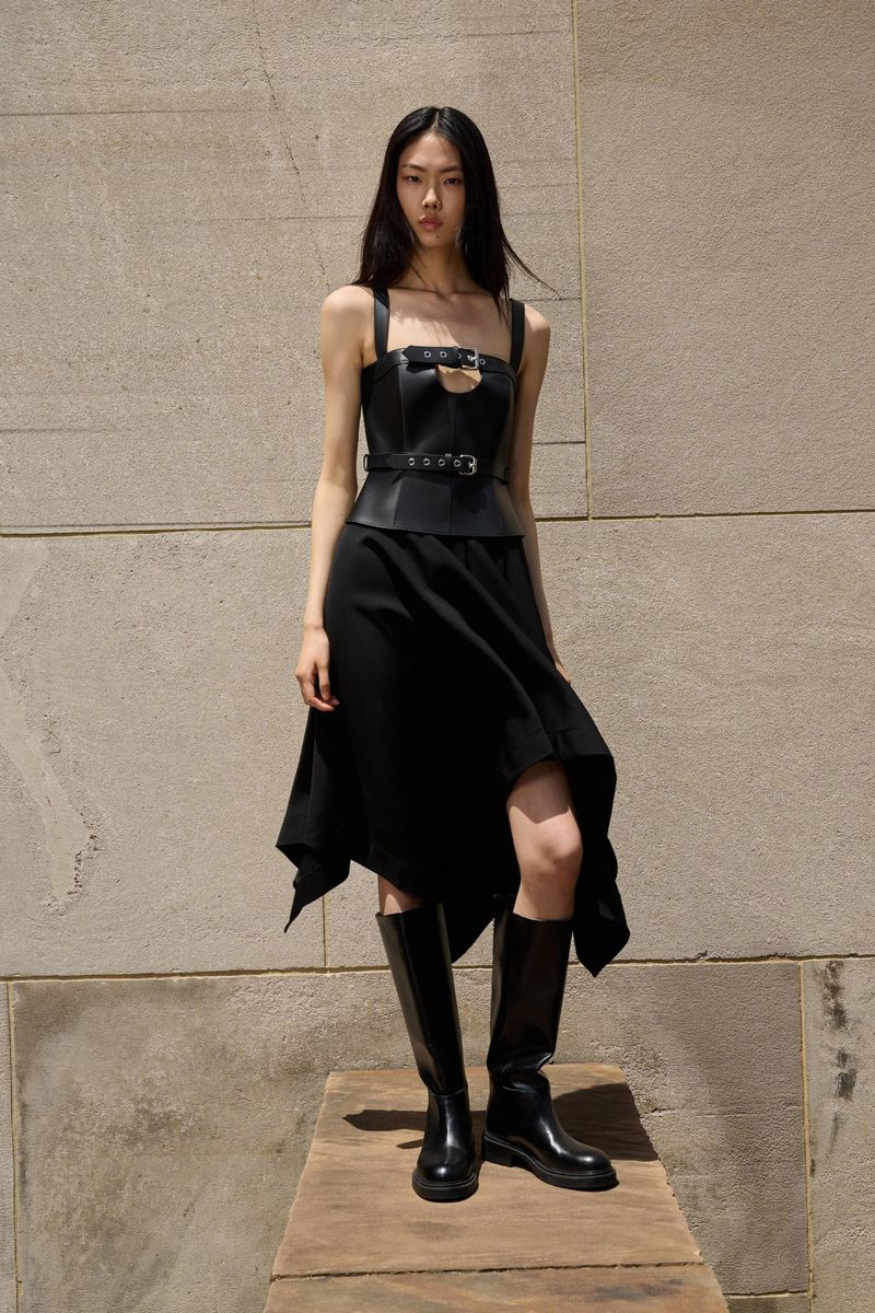 An Asian woman in a black dress with a leather bodice and black boots stands in front of a textured wall on a wooden platform.