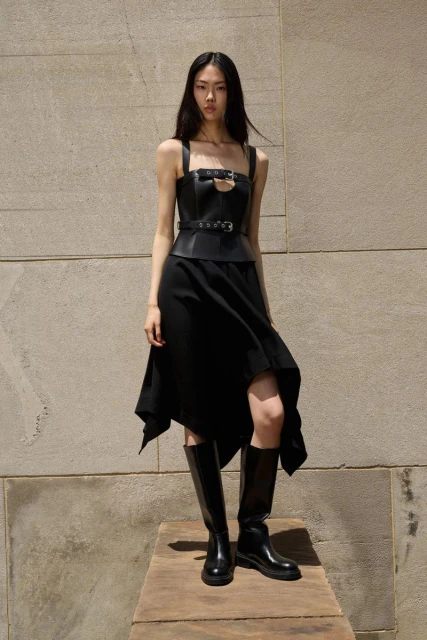 An Asian woman in a black dress with a leather bodice and black boots stands in front of a textured wall on a wooden platform.