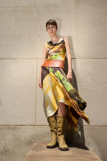 A slender woman with short brown hair stands against a textured wall wearing a print top, draped skirt, wide brown belt, and knee-high boots.