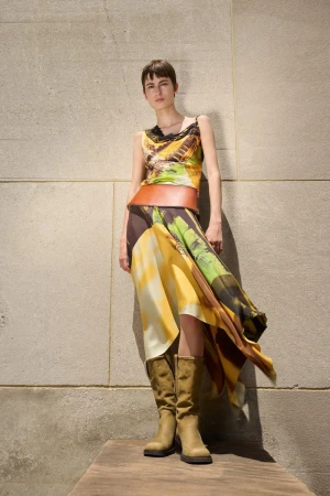 A slender woman with short brown hair stands against a textured wall wearing a print top, draped skirt, wide brown belt, and knee-high boots.