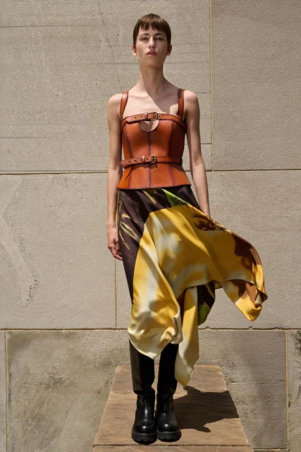 Fashion model poses in brown leather corset top and floral print skirt with knee-high boots against a neutral textured wall.
