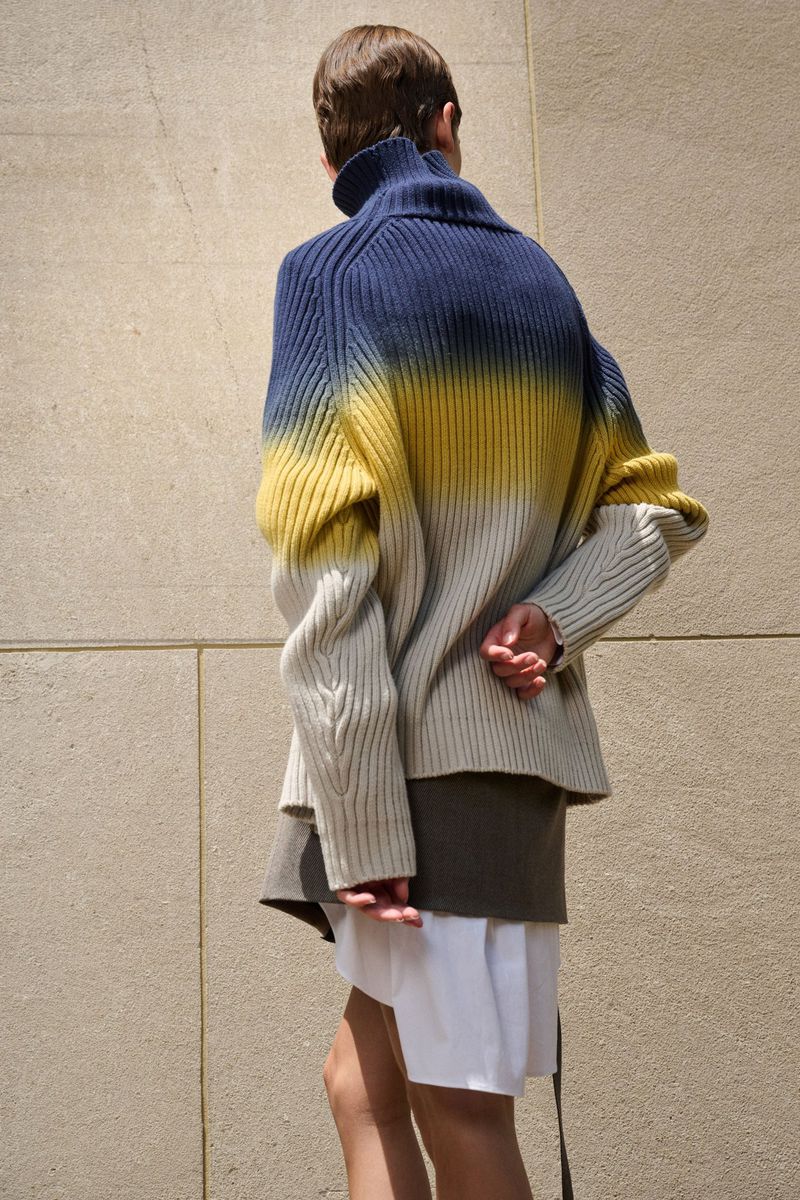 Back view of a person wearing a blue, yellow, and white ombre knit sweater and layered skirts standing against a neutral wall.
