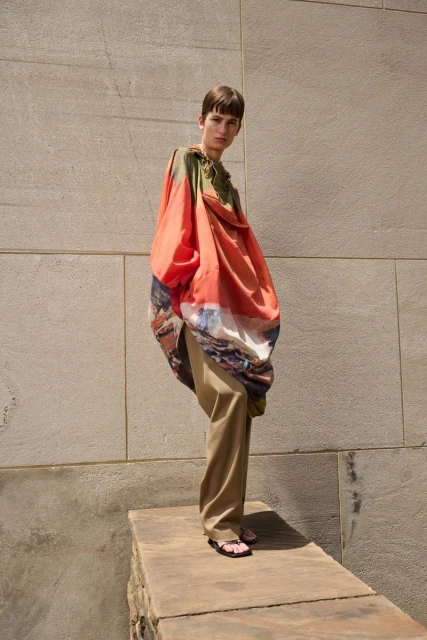 Fashion shot of a person wearing orange painterly design, posing on a stone structure with a concrete wall background in bright daylight.