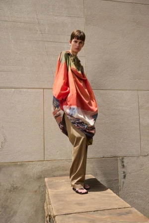 Fashion shot of a person wearing orange painterly design, posing on a stone structure with a concrete wall background in bright daylight.