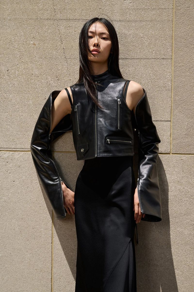 An Asian adult female poses in a black skirt and black leather jacket with detached sleeves, against a neutral-toned wall backdrop.