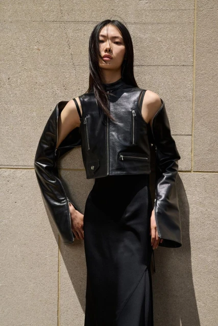 An Asian adult female poses in a black skirt and black leather jacket with detached sleeves, against a neutral-toned wall backdrop.