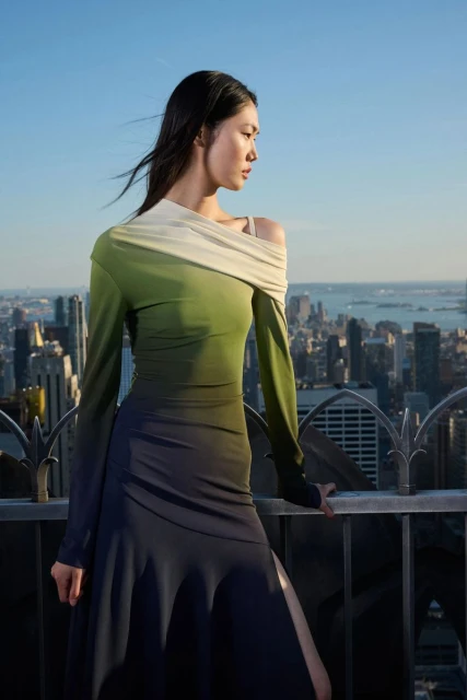Asian woman poses in a long-sleeve ombre dress on a rooftop with a city skyline background under a clear, blue sky.