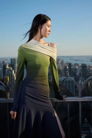 Asian woman poses in a long-sleeve ombre dress on a rooftop with a city skyline background under a clear, blue sky.