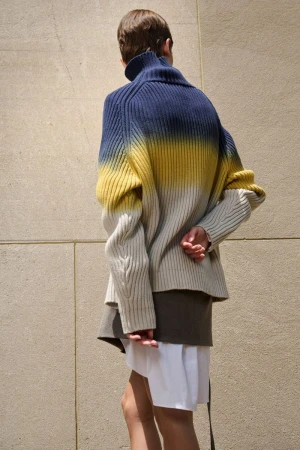 Model seen from behind wearing an ombre navy, yellow, and cream ribbed knit turtleneck sweater against a beige stone wall.