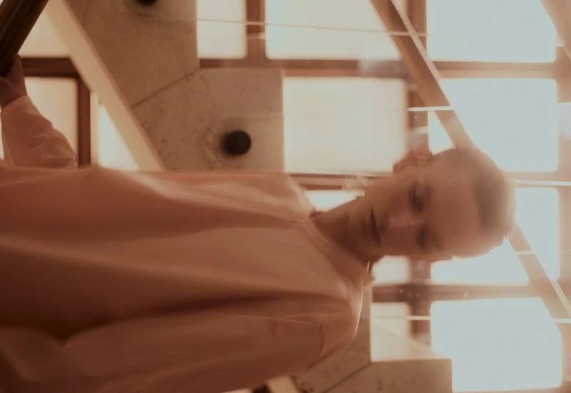 Young woman descends stairs indoors bathed in bright natural light. The photo has a soft focus with geometric elements in the background.