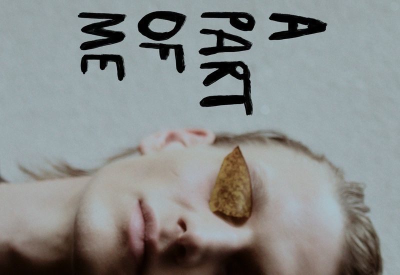 A person with fair skin laying down with leaf covering one eye next to the words 'A Part of Me' in handwritten text.