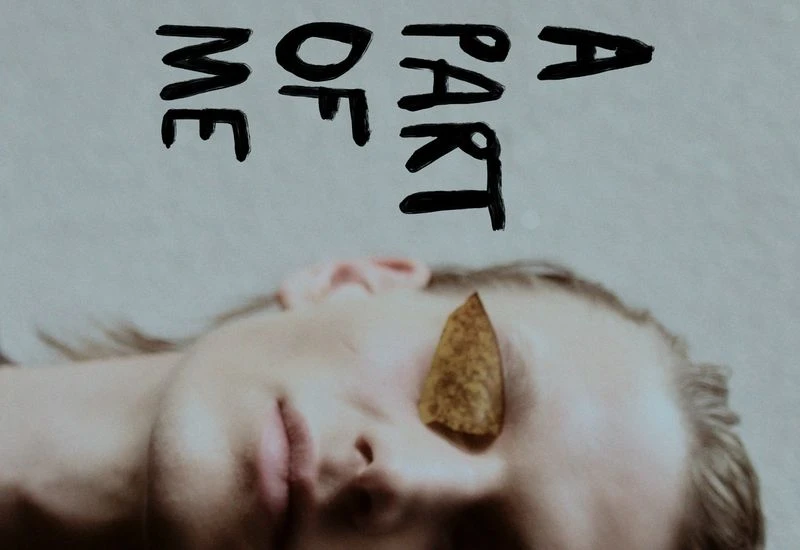 A person with fair skin laying down with leaf covering one eye next to the words 'A Part of Me' in handwritten text.