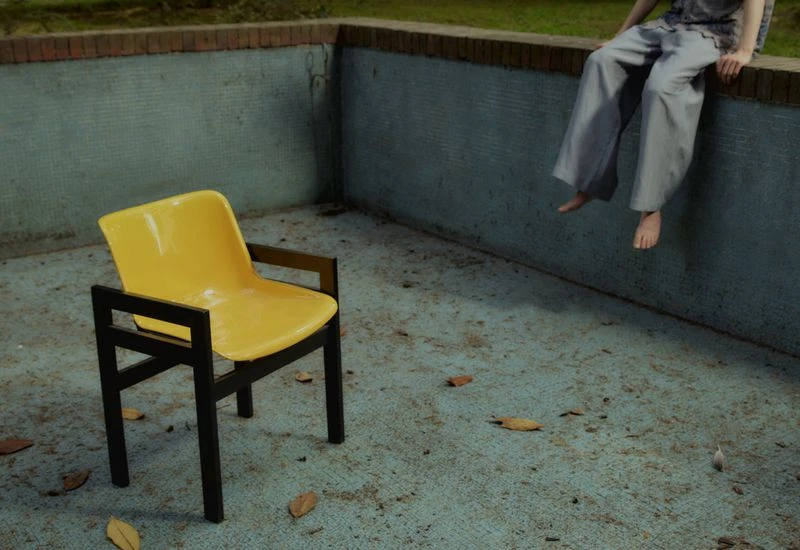 Person sits on wall near a yellow chair. The image has cool tones, casual clothing, and an outdoor setting with some visible leaves.