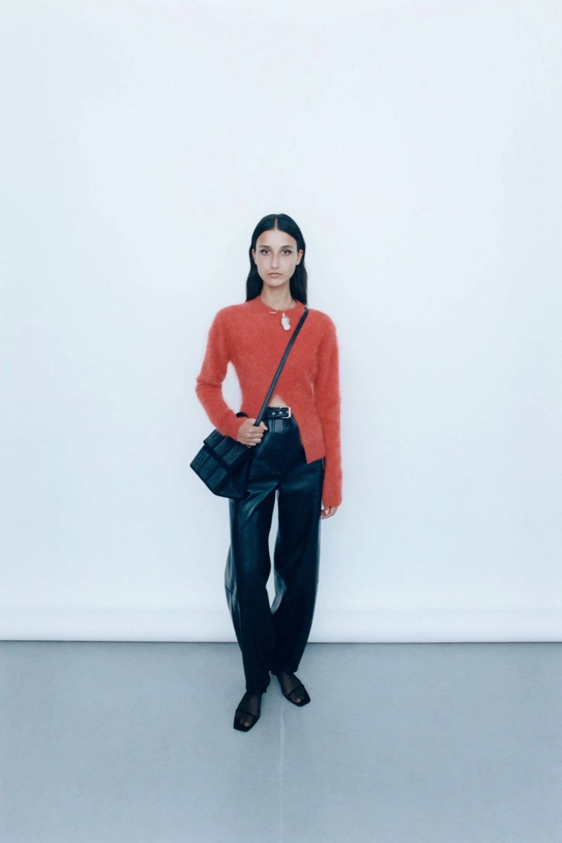 A woman in an orange sweater and black leather pants poses against a white background, wearing a black cross-body bag and black flats.