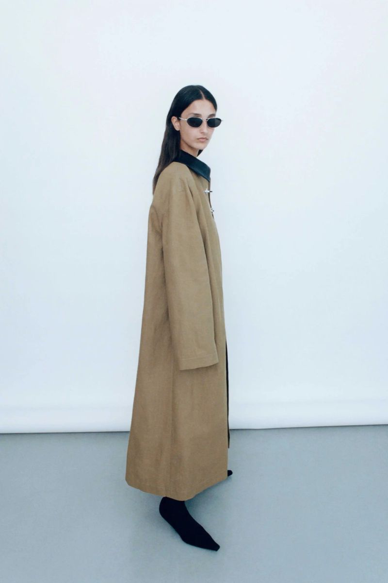 A side-view studio shot of an adult woman posing in a tan overcoat, black top, black socks and dark sunglasses against a white backdrop.