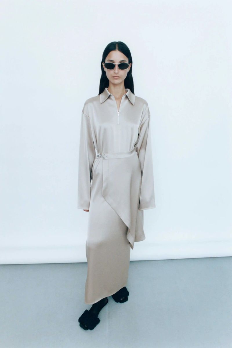 A woman wearing a beige dress and black sunglasses stands against a white background in a fashion editorial-style full shot.