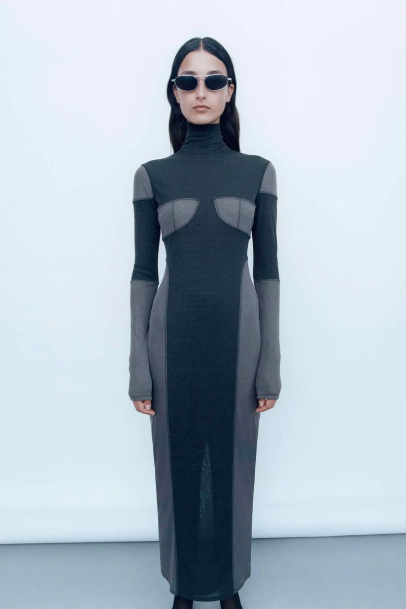 Woman in a dark two-tone turtleneck maxi dress and sunglasses stands in a bright studio against a neutral white background.